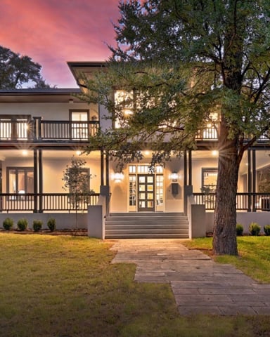 Modern farmhouse with white exterior, black railings, and wraparound porches at dusk with mature trees and manicured lawn.