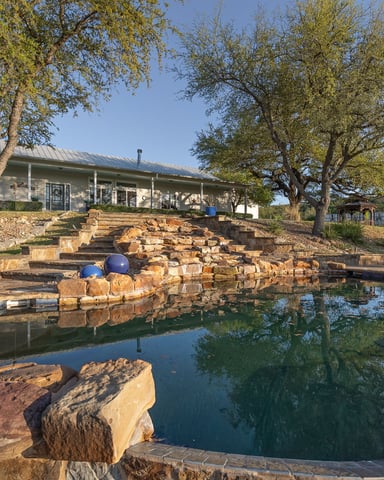 Modern ranch house with metal roof overlooking stone-lined pond and waterfall, surrounded by mature trees and seating areas