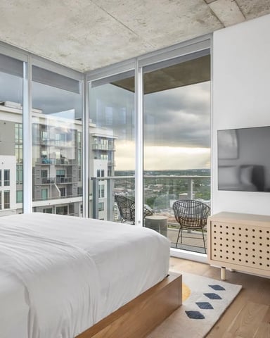 High-rise bedroom with white bed, wooden dresser, wall-mounted TV, and floor-to-ceiling windows overlooking city skyline and balcony.