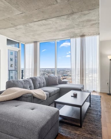 Modern high-rise living room with gray sectional sofa, concrete ceiling, floor-to-ceiling windows overlooking cityscape, and green accent wall.