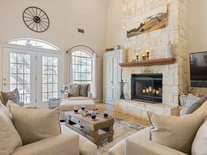 Spacious living room with stone fireplace, cream armchairs, wooden coffee table, and French doors opening to snowy landscape.
