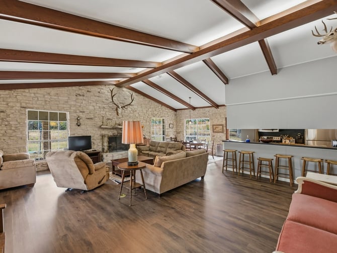 Open-plan living room with exposed wood beams, stone fireplace, tan upholstered seating, and kitchen with bar seating