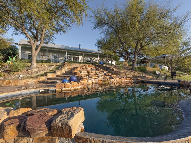 Modern ranch house with metal roof overlooking a natural stone pond and waterfall feature, surrounded by mature trees and seating areas.